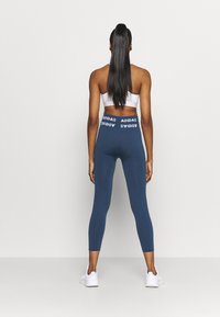 Leggings azules con una textura acanalada y el logo blanco de "ADIDAS" en la cinturilla, combinados con un sujetador deportivo blanco y zapatillas blancas.