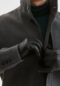 Grauer Fischgrätenmantel mit hohem Kragen, schwarzen Knopfverschlüssen und maßgeschneiderten Ärmeln; schwarze Lederhandschuhe greifen die Öffnung des Mantels.