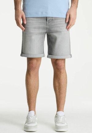 Mees kannab heledaid halli värvi ülesrullitud teksapükse, valgeid tossusid ja helesinist särki, seistes ühevärvilise tausta ees.