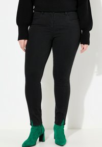 Jeans negros ajustados con un diseño entallado, cintura alta y slit en los laterales del dobladillo, combinados con botines de ante verde de tacón alto.