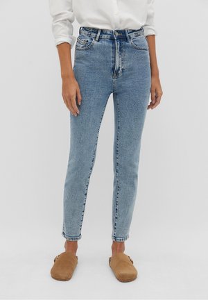 Personne portant un jean skinny taille haute bleu clair, une chemise blanche à boutons avec manches retroussées et des chaussures marron sans lacets sur fond uni.
