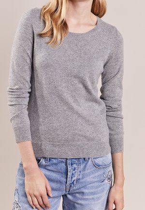 Kvinde iført en lysegrå langærmet sweater og blå jeans med blomsterbroderi, stående foran en ensfarvet baggrund.