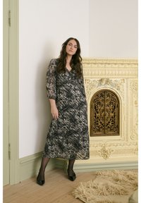 Femme en robe noire à motifs et talons noirs se tenant à côté d'une cheminée vintage ornée de couleur crème dans une pièce avec des garnitures vert clair.