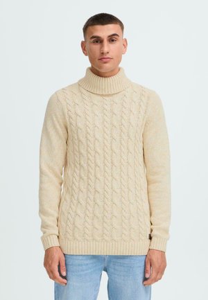 Beige, grob gestrickter Rollkragenpullover mit gerippten Bündchen und Saum. Besitzt ein strukturiertes Muster und eine taillierte Passform. Getragen mit hellblauen Jeans.
