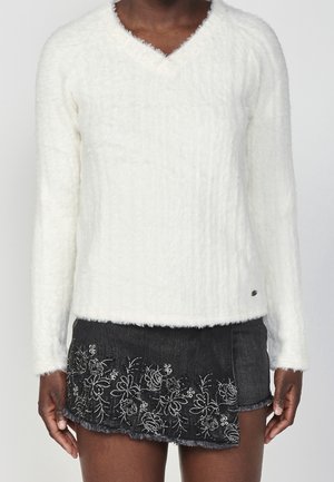 Osoba o ciemnej karnacji ubrana w białą, puszystą bluzę z dekoltem w serek oraz czarną mini spódniczkę dżinsową z białą kwiatową haftą.