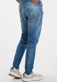 Blauwe denim jeans met een slim fit, voorzien van achterzakken en een bruine leren patch op de tailleband. Draagt met grijze sport sneakers.