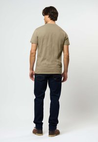 Beige puuvillane t-särk ümarate kraega, lühikeste varrukatega, kombineeritud tumesiniste teksade ja pruunide saabastega, vaadatud tagant.