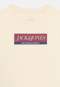 Kremfarget T-skjorte med en sentrert rektangulær logo som leser "JACK & JONES" over teksten "SINCE NINETEEN NINETY."