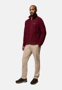Giacca in pile marrone con zip frontale e tasche laterali, abbinata a pantaloni beige e scarpe da trekking marroni, con tessuto testurizzato.