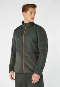 Giacca in pile verde con una parte superiore liscia, dotata di cerniera arancione e dettagli arancioni. Tessuto testurizzato e morbido nella parte inferiore e nelle maniche.