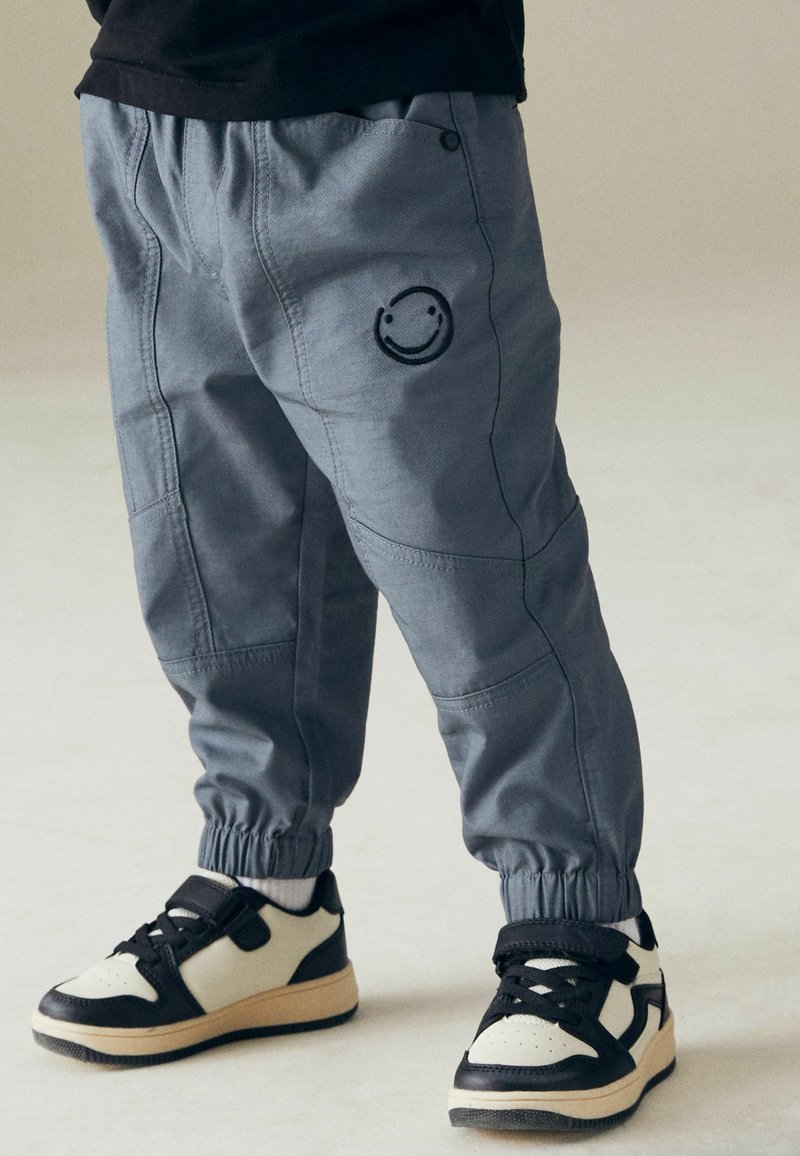 Enfant portant un pantalon de jogging gris avec un smiley cousu sur la cuisse et des baskets noir et blanc, debout sur un sol clair.