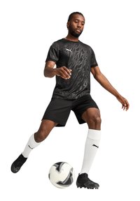 Schwarzes sportliches T-Shirt mit strukturiertem Muster, schwarze Shorts, weiße Fußballsocken und schwarze Fußballschuhe; Spieler interagiert mit einem Fußball.