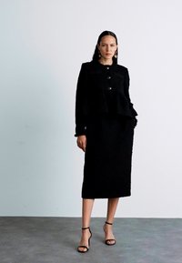 Black two-piece dress featuring a structured peplum jacket with button details and a long skirt. Smooth texture and form-fitting silhouette.