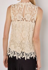 Haut en dentelle beige avec motifs floraux, col montant et dos avec ouverture en forme de goutte. Comprend des détails en crochet délicats et un ourlet festonné.