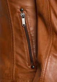 Veste en cuir marron avec une poche zippée visible, dotée d'une fermeture éclair noire et d'un bouton-pression. Texture lisse et lignes de design ajustées.