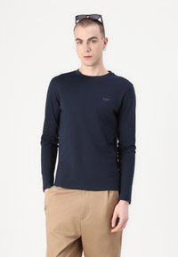 Maglietta a maniche lunghe color navy con scollo a giro e piccolo logo sul petto, abbinata a pantaloni beige. Texture liscia e design aderente.