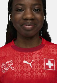 Punane spordijersey lühikeste varrukatega, millel on valge Puma logo, Šveitsi lipu klapp ja mustriga tekstuur, millel on risti detailid.