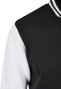 Black and white varsity jacket featuring a black body, white sleeves, and a striped collar. Made from a smooth, flexible fabric.