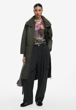 Long olive green quilted coat over a patterned top featuring purple and green hues, paired with loose black trousers and black shoes.