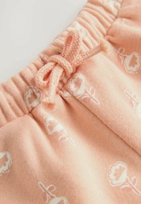 Mjukt rosa tyg med blommönster, med en flätad knutdetalj och elastisk midja för justerbar passform. Texturen verkar slät.