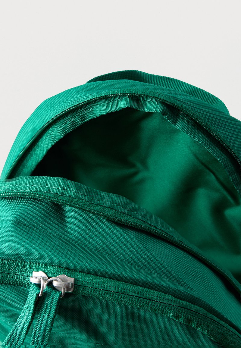 Grüner Stoff-Rucksack mit teilweise geöffnetem Hauptfach und vorderer Reißverschlusstasche vor weißem Hintergrund.