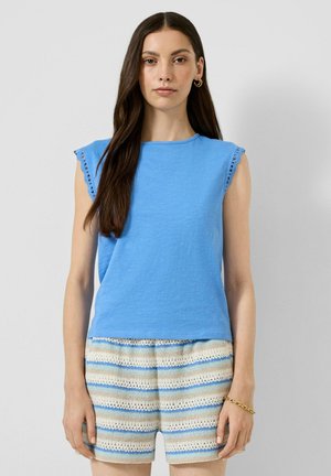 Femme aux longs cheveux foncés portant un top bleu sans manches avec bordure ajourée et un short tricoté avec des rayures beige, bleu et blanc, debout sur un fond gris.