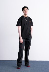 Zwarte korte mouwen t-shirt met logo, gecombineerd met zwarte broeken met rode stiksels. Draagt zwarte schoenen en een zilveren ketting. Effen achtergrond.