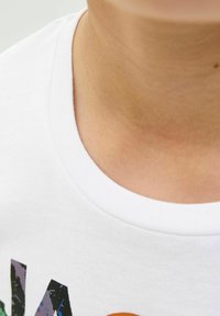 T-shirt bianco in cotone con scollatura tonda; cucitura del colletto visibile e una piccola sezione di grafiche stampate colorate vicino al bordo inferiore.