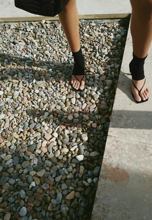 Persona con sandalias negras de calcetín, de pie con un pie sobre guijarros de piedra y el otro en un suelo de baldosas bajo la luz del sol.