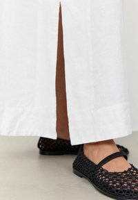 Witte linnen maxi-jurk met een zijsplit, gecombineerd met zwarte geweven flats met een band over de voet. Soepele textuur en een minimalistisch ontwerp.