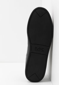 Semelle de chaussure en caoutchouc noir avec une surface texturée, un motif triangulaire et un logo "MK" en relief. Bords lisses, design minimaliste.