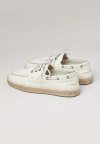 Zapatos de barco de cuero blanco con punta redonda, cordones grises, ojales metálicos y suela de esparto trenzada. Presentan un sutil logo en el lateral.