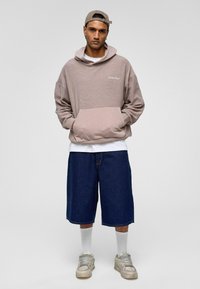 Overmaatse beige hoodie met een voorzak en wit logo, gecombineerd met denim shorts tot de knie en grijze sneakers, geaccentueerd met witte sokken.
