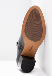 Botín de cuero negro con una suela texturizada, que cuenta con una capa de caucho natural y un tacón cuadrado. Logo "Clarks" visible cerca del tacón.