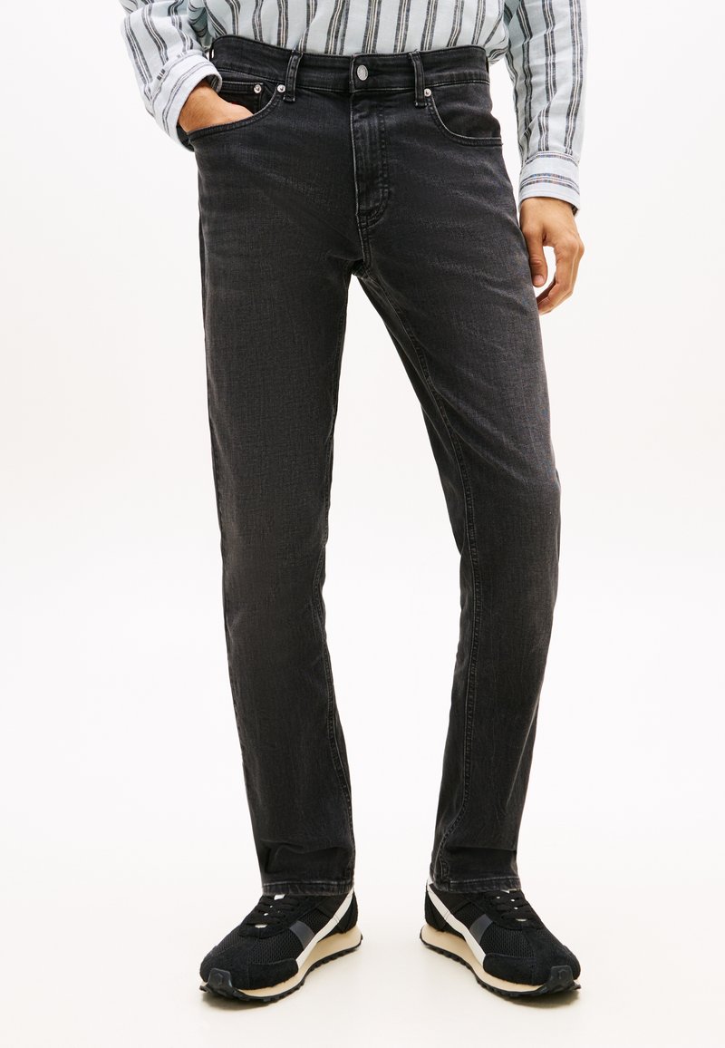 Άνδρας που φοράει μαύρο στενό τζιν παντελόνι, μαύρα και λευκά αθλητικά παπούτσια και ριγωτό μακρυμάνικο πουκάμισο, με το ένα χέρι στην τσέπη.