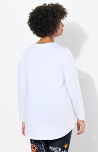Chemise blanche à manches longues avec un col arrondi et une coupe décontractée, présentant une texture lisse et un ourlet incurvé.