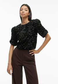 Melnas pailletes virskrekls ar pūstām piedurknēm, ribota tekstūra, apaļa kakla līnija, kombinēts ar augsta vidukļa brūnām biksēm. Gluda auduma un pieguloša dizaina.