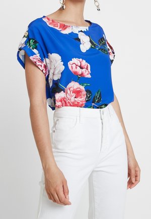 Blusa corta de mangas en un vibrante azul, con flores rosas y blancas, de textura suave. Combinada con pantalones vaqueros de cintura alta blancos.