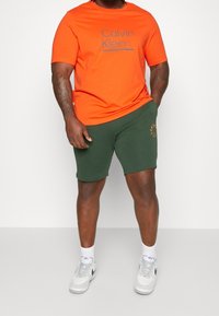 Pomarańczowa bawełniana koszulka z szarym tekstem logo oraz zielone bawełniane szorty z małą pomarańczową grafiką na lewej nogawce. Białe sneakersy.