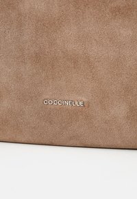 Braune Wildlederhandtasche mit glatter Oberfläche und einem silbernen "Goccinelle"-Logo auf der Vorderseite. Schlichtes Design und klare Linien.
