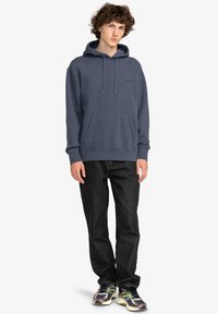 Sudadera azul marino con cordones, bolsillo frontal y puños de canalé. Combinada con jeans negros y deportivas multicolor. Ajuste casual.