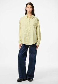 Langarmige gele blouse met witte en groene strepen, gecombineerd met donkerblauwe wijduitlopende jeans en zwarte schoenen.