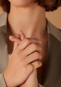 Goldring mit einem dekorativen Mittelpunkt, der wie ein Edelstein geformt ist, getragen an einer Hand mit gepflegten Nägeln, vor einem neutralen Hintergrund.