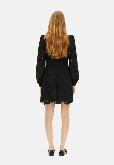 Frau mit langen roten Haaren, die ein schwarzes Blumenspitzenkleid mit langen Puffärmeln und schwarzen flachen Schuhen trägt, steht mit dem Rücken zugewandt.