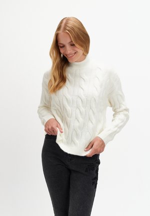 Biały sweter z dzianiny w warkocze z wysokim kołnierzem i prążkowanymi mankietami, zestawiony z czarnymi dżinsami z kwiatowym haftem na udzie.