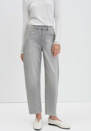 Personne portant un jean large gris clair, un haut blanc à manches longues et des chaussures plates blanches à enfiler, se tenant devant un fond uni.