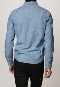 Vaaleansininen denimpaita, jossa on nappikiinnitteinen kaulus, pitkät hihat ja yhden napin kalvosimet, teksturoidusta kankaasta ja istuvalla leikkauksella.