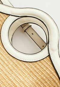 Gros plan d’un sac en paille tressée avec une anse circulaire en cuir blanc et des coutures visibles sur un fond uni.