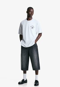Bílý bavlněný tričko s černým grafickým potiskem, spojený s tmavými džínovými culottes. Modelka má na sobě bílé ponožky a černé mokasíny.