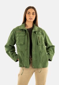 Chaqueta de algodón verde con múltiples bolsillos frontales, cierres con cremallera y a presión, dobladillo ajustable con cordón y un sutil bordado en el cuello.